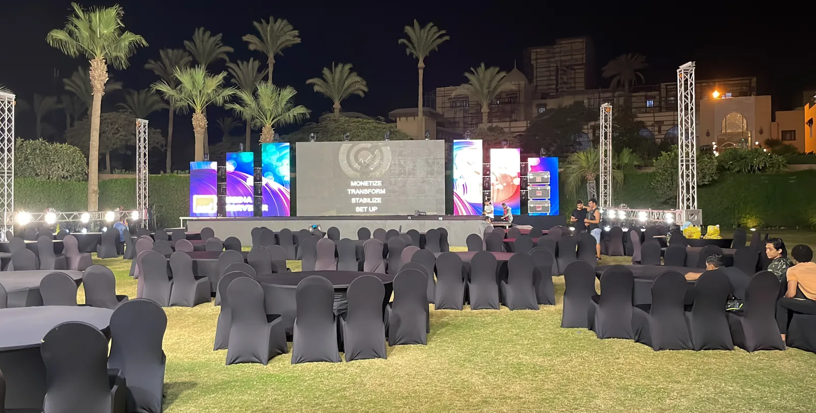 The lawn at night: rows of black-covered chairs and tables facing a lit stage of screens between truss towers.