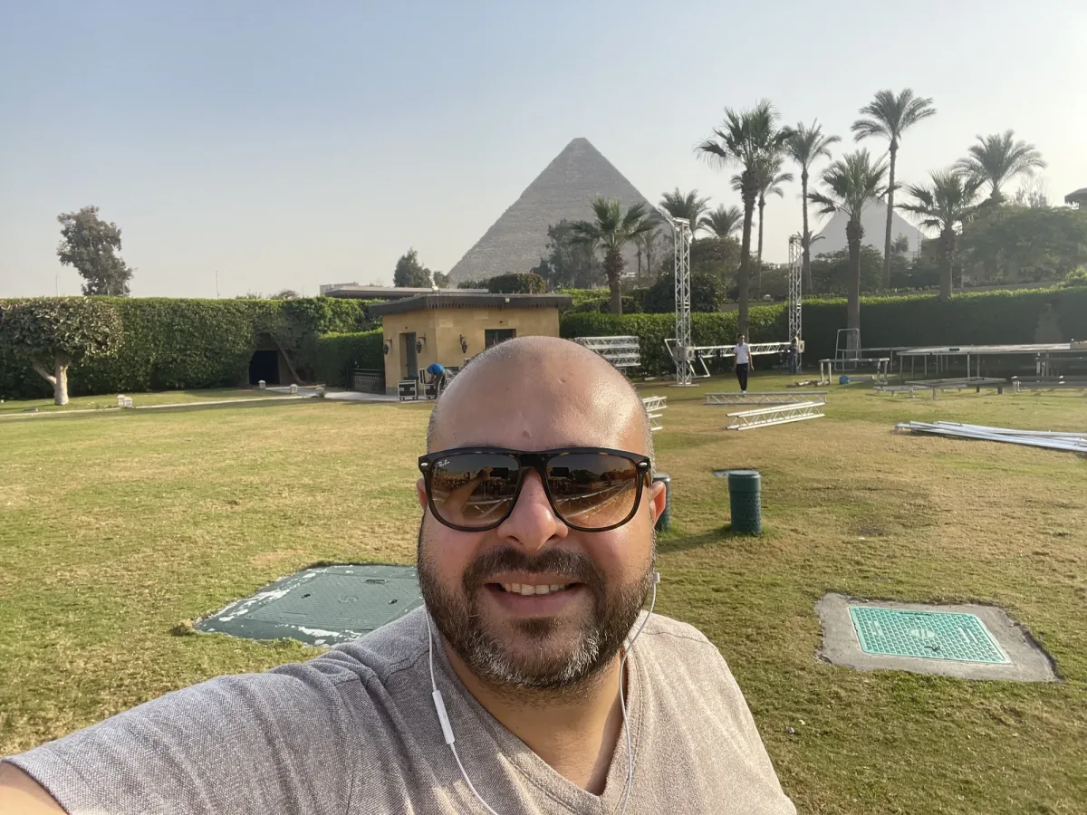 Antoni Gerges smiling in sunglasses on the lawn, truss towers and the Great Pyramid behind him.
