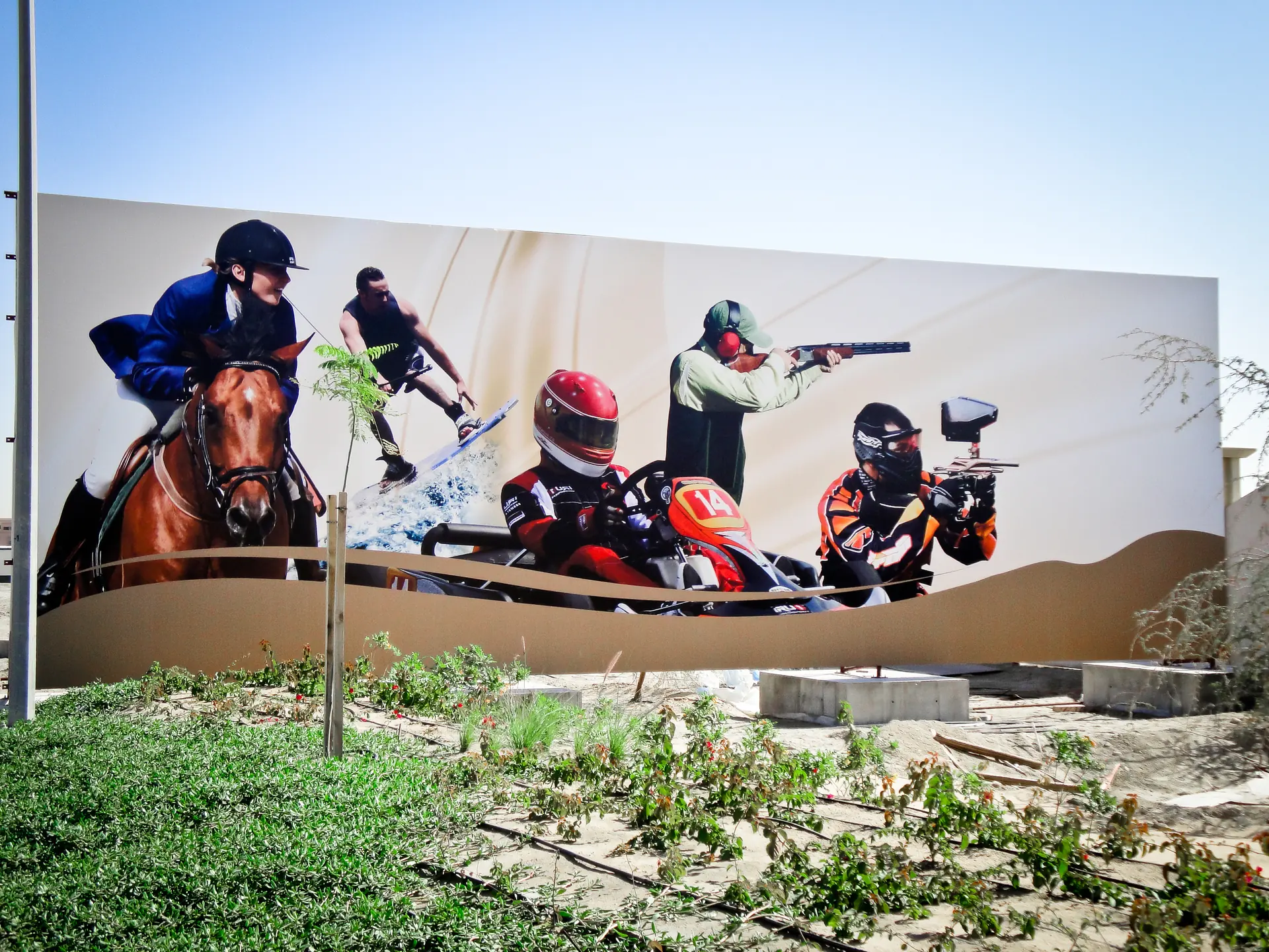 Photograph: a hoarding on the building site showing the rider, the wakeboarder, the kart driver, the shooter and the paintballer above a brown wave