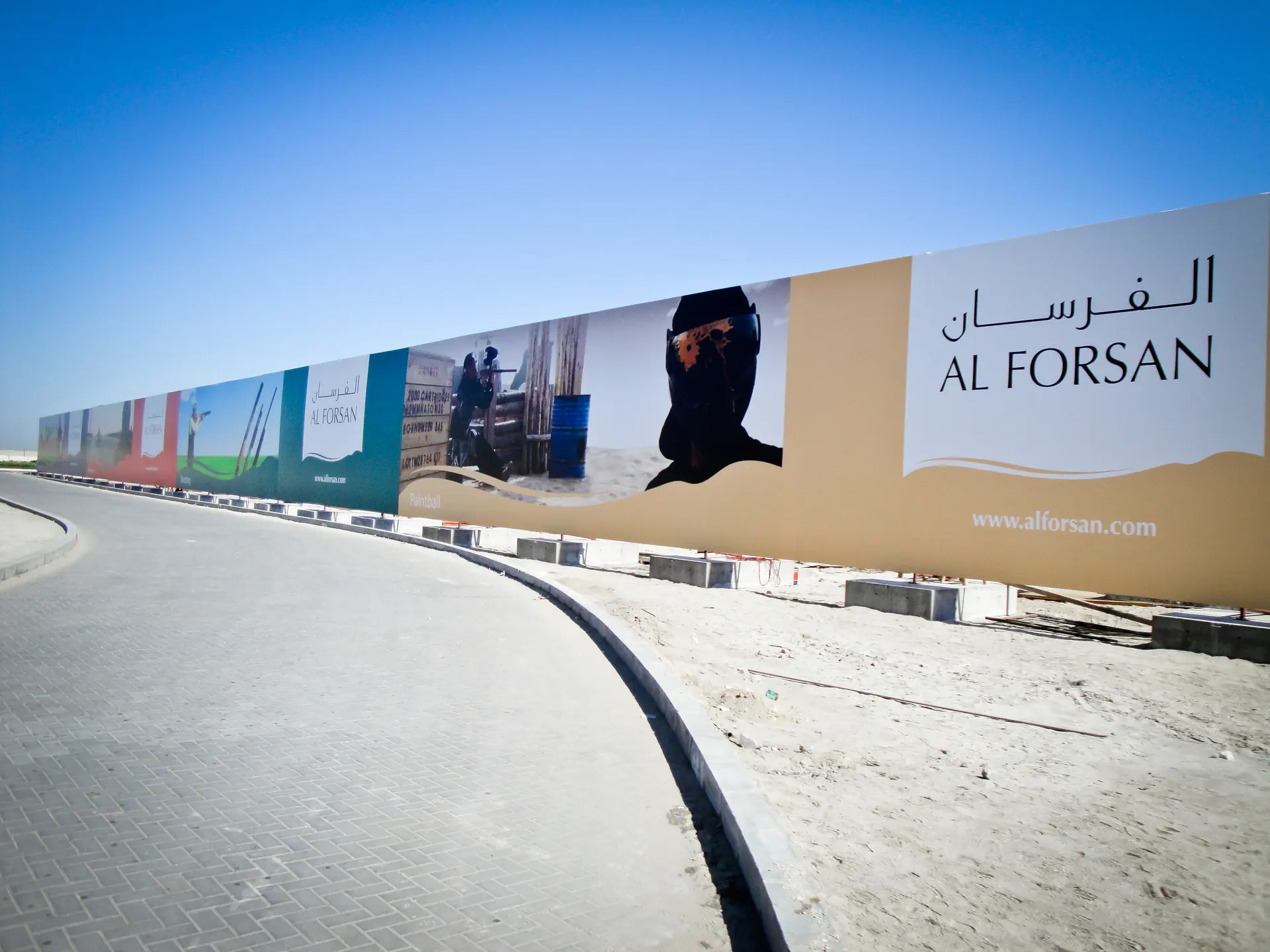 Photograph: a long hoarding following a curved road in the sand, with the two names of the resort on its first panel
