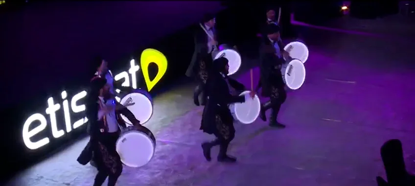 A frame of the event film: drummers with lit drums walk past giant lit letters, etisalat, on the sand